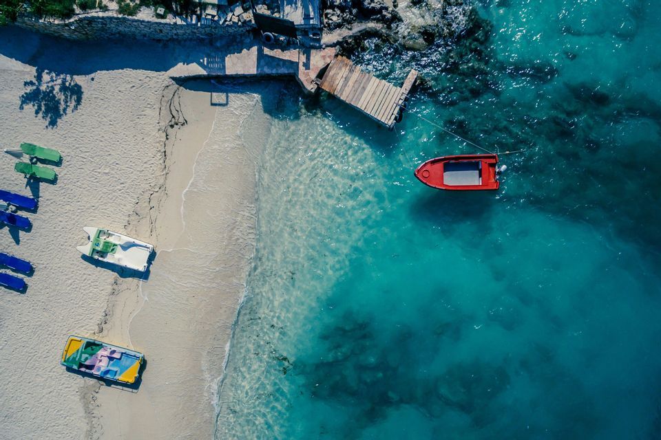 Top-down aerial view of a sandy beach with several boats on the sand and a red boat floating in turquoise water by a dock.