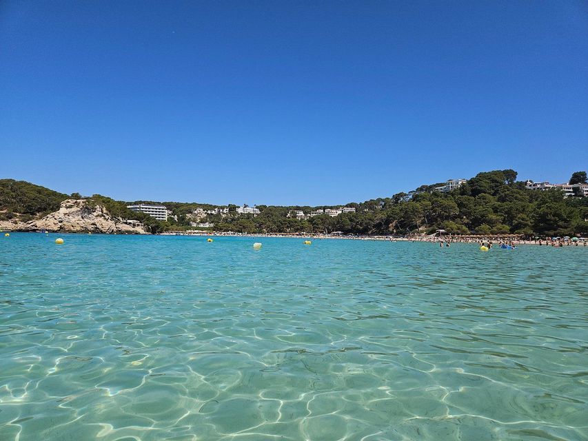 Vista dalle acque limpide e turchesi di una cala, verso una spiaggia sabbiosa affollata e verdi colline alberate.