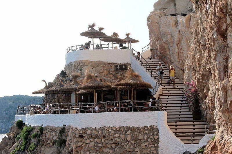 Un ristorante a più livelli con ombrelloni di paglia, incastonato nella parete di una scogliera rocciosa e affiancato da una lunga scalinata.