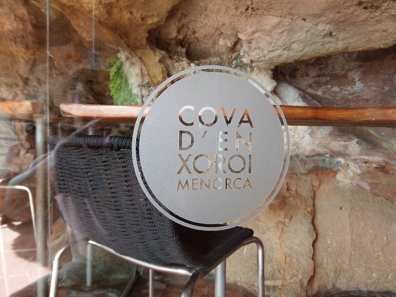 Un logo circolare smerigliato di Cova d'en Xoroi Minorca su una porta a vetri, con l'interno simile a una grotta e una sedia nera visibili.
