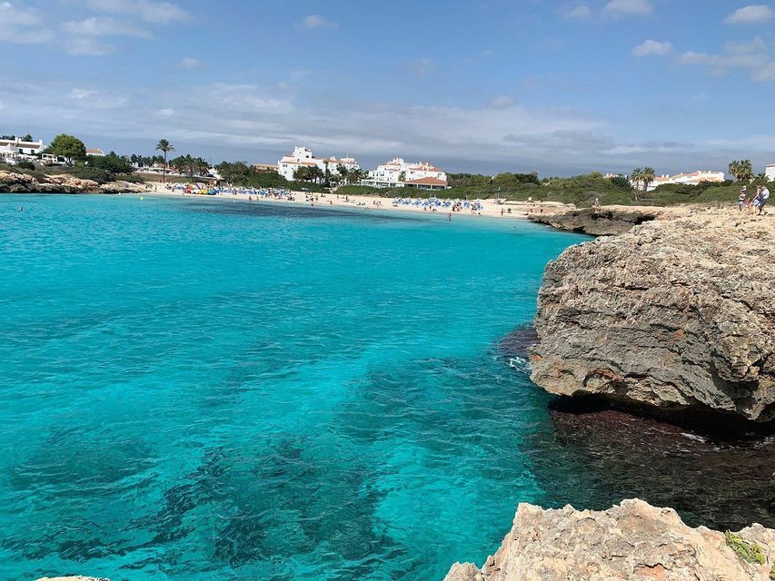 Una vista da una costa rocciosa su una cala con acqua turchese limpida, una spiaggia sabbiosa e edifici bianchi sullo sfondo.