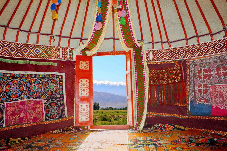 Vue de l'intérieur d'une yourte colorée, regardant à travers une porte rouge ouverte sur une vallée verdoyante et des montagnes enneigées.