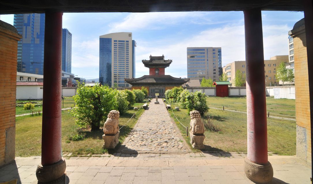 Un temple asiatique traditionnel avec un chemin de pierre et des jardins, encadré de piliers rouges, avec des gratte-ciel modernes en arrière-plan.