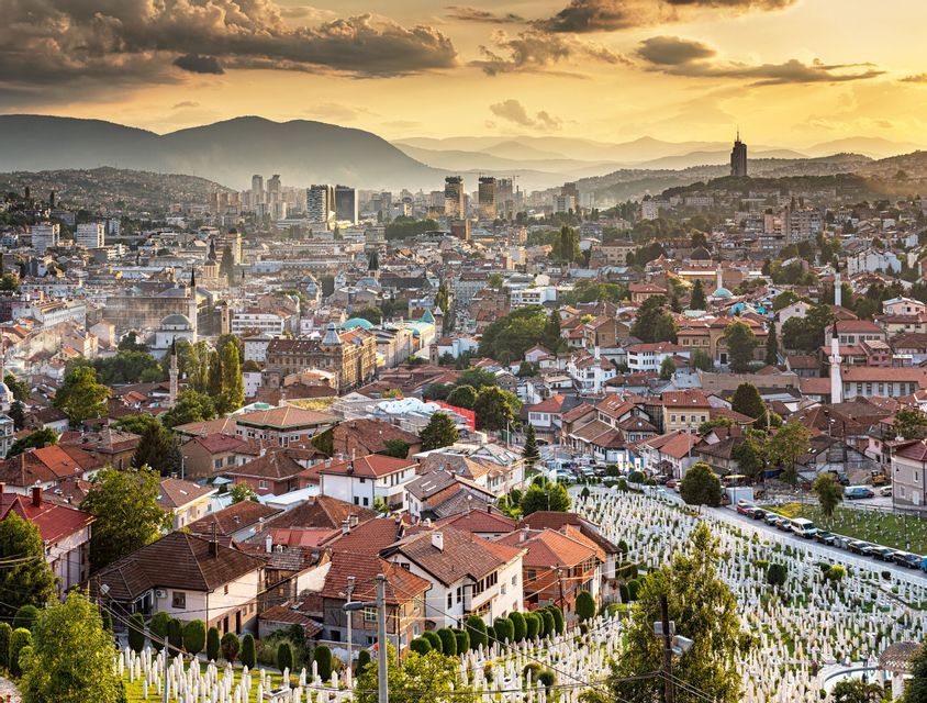 Ein erhöhter Blick auf eine weitläufige Stadtlandschaft mit rotgedeckten Gebäuden und einem großen Friedhof im Vordergrund, vor einer Bergkulisse bei Sonnenuntergang.