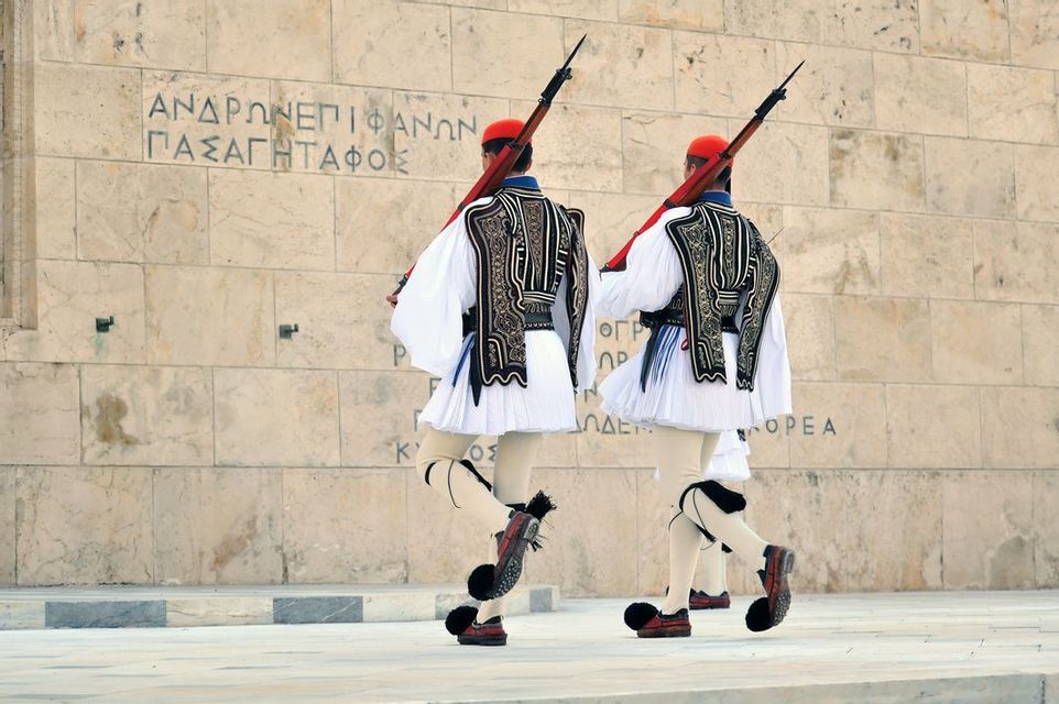 Due guardie in uniformi tradizionali greche con fucili e scarpe con pon-pon marciano all'unisono davanti a un muro di pietra con iscrizioni.