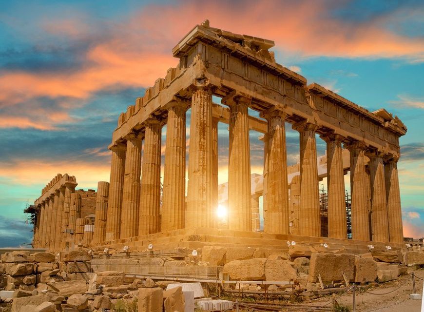 Le rovine di un antico tempio con grandi colonne sono illuminate dal sole al tramonto su un cielo colorato.
