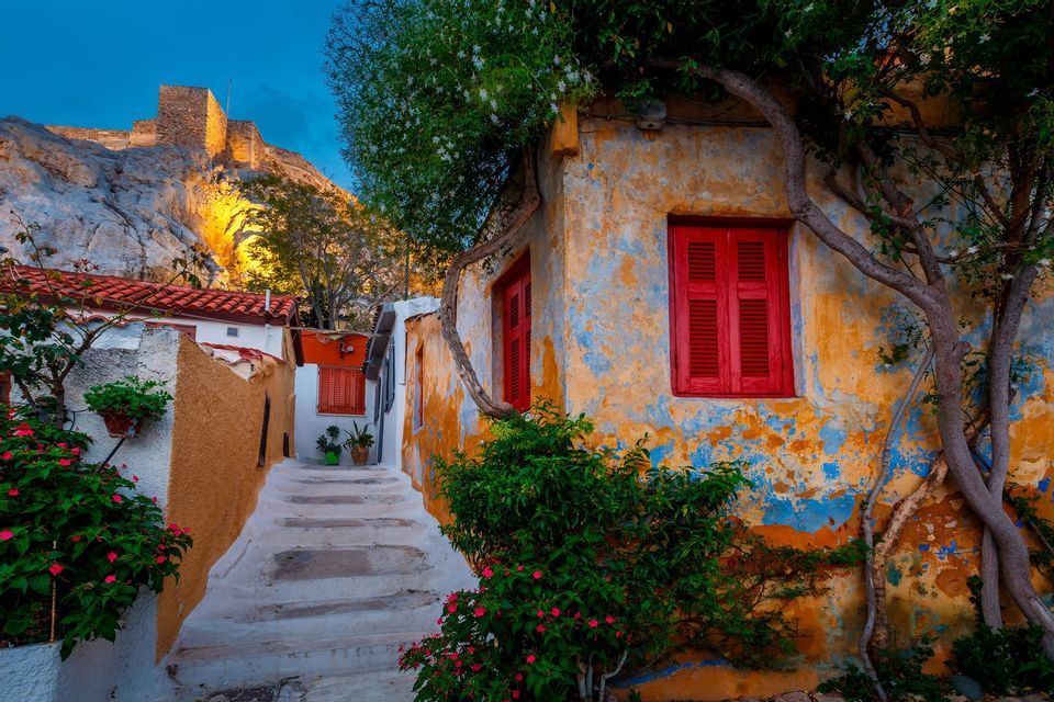 Un vicolo in pietra si snoda accanto a un edificio rustico con persiane rosse. Sullo sfondo, una fortezza si erge su una collina rocciosa illuminata.
