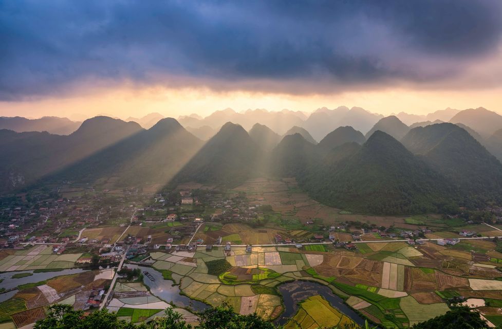 Des rayons de soleil percent les nuages sombres sur une vallée de montagnes vertes, des rizières en patchwork et un petit village.
