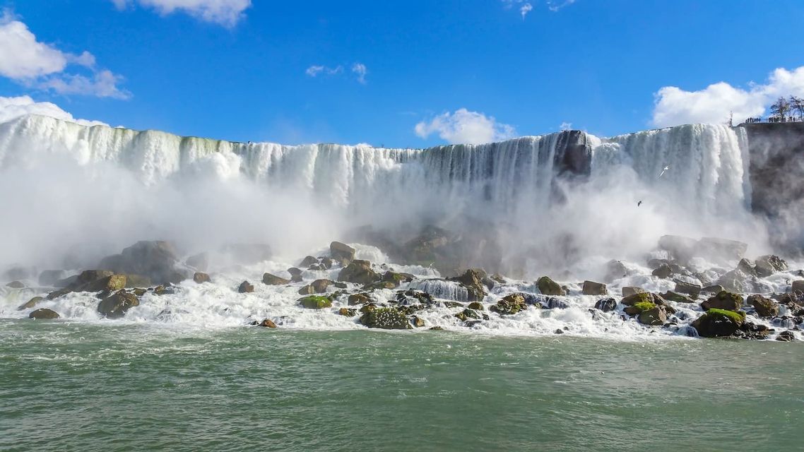 Una cascata ampia e potente si getta da una scogliera su grandi rocce sottostanti, creando una fitta nebbia sotto un cielo blu.