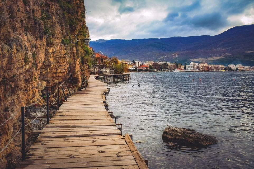 Una passerella di legno si estende lungo una scogliera rocciosa accanto a un lago, con una città e montagne in lontananza sotto un cielo nuvoloso.