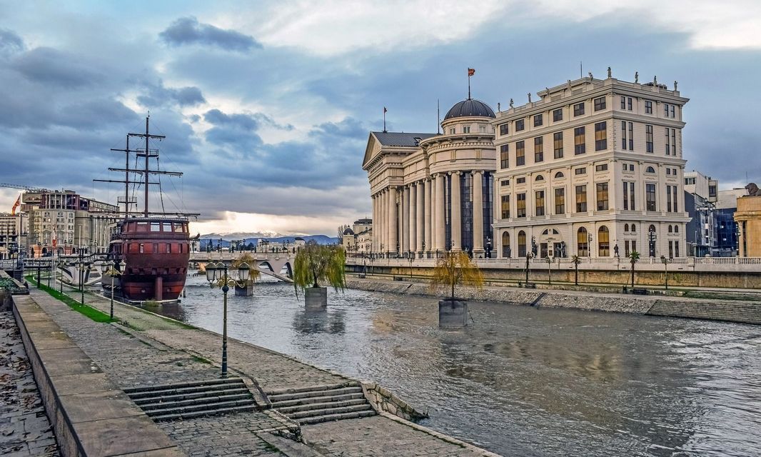Una vista lungo il fiume di una città con una nave di legno attraccata accanto a un grande edificio neoclassico sotto un cielo nuvoloso.