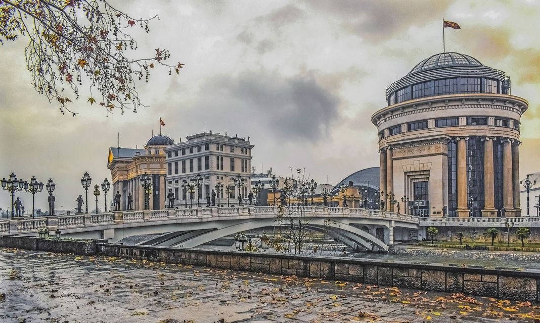 Edifici neoclassici ornati e un ponte fiancheggiato da statue si ergono su un fiume in una giornata autunnale nuvolosa con foglie a terra.