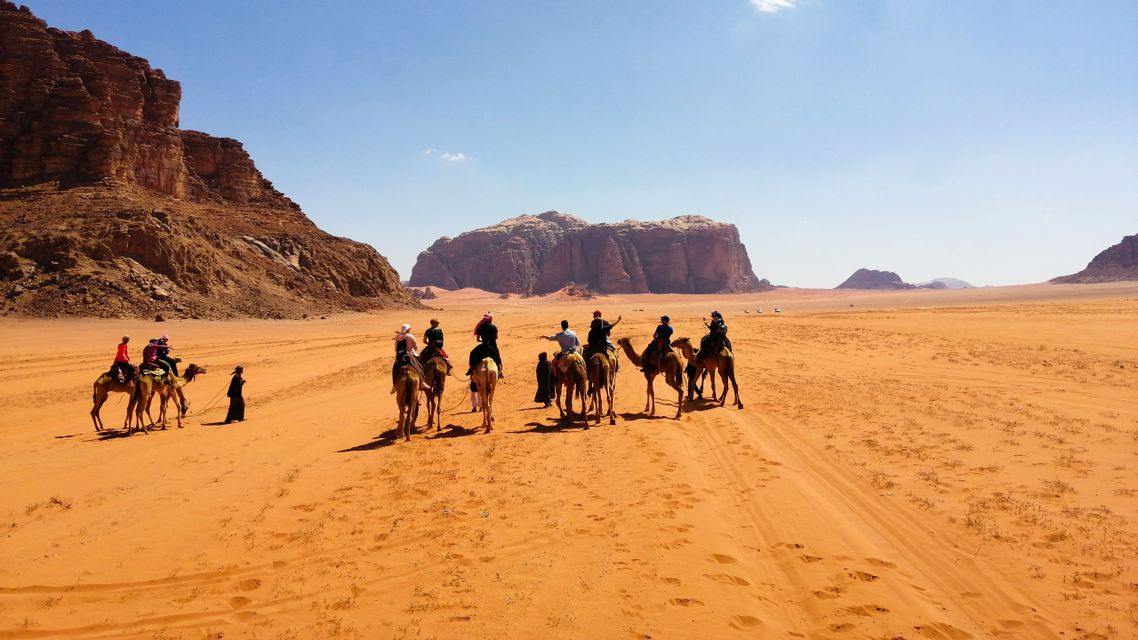 A WeRoad group trip rides camels in a line through a red sand desert, with rocky mountains in the background.