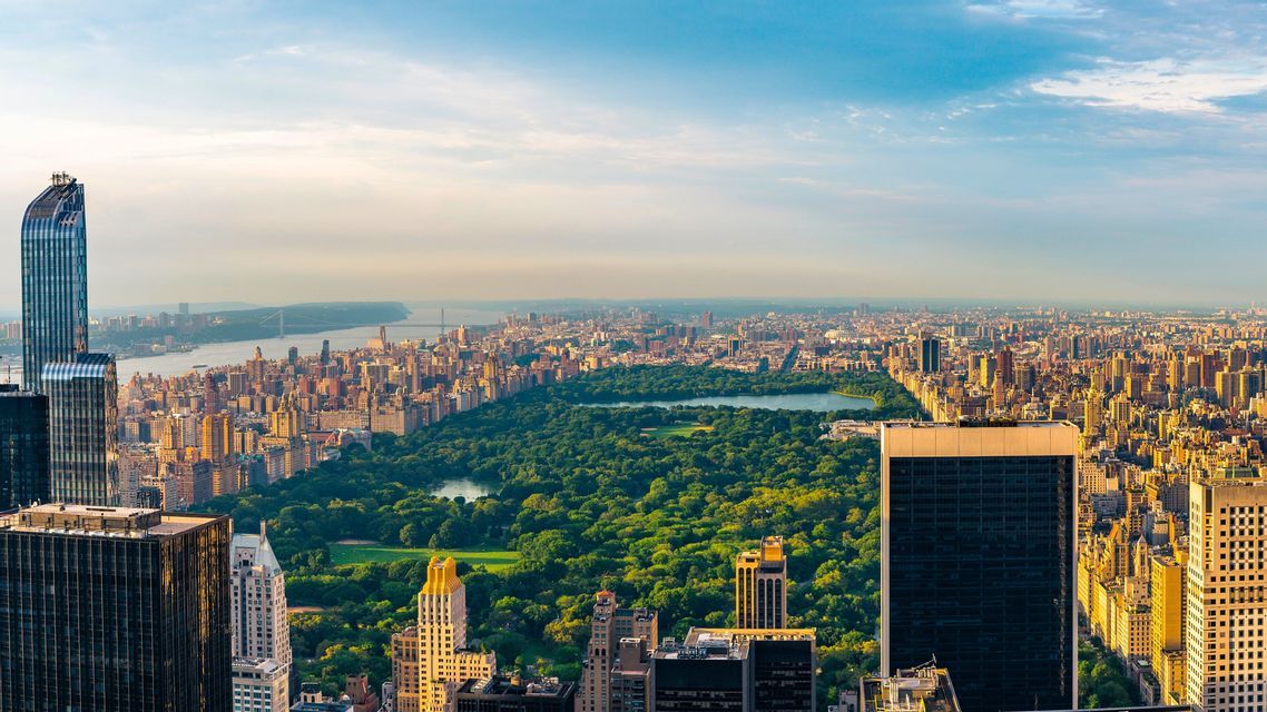 An aerial view of a sprawling cityscape with skyscrapers surrounding a large, green central park and a river in the distance.