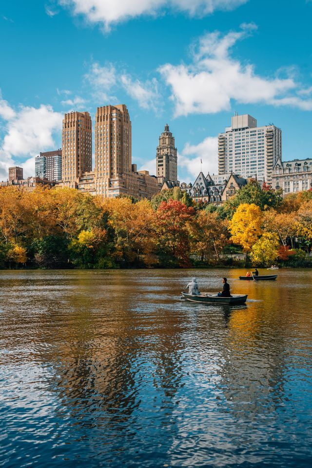 Persone su barche a remi su un lago con uno skyline cittadino visibile dietro una fila di alberi con fogliame autunnale colorato.