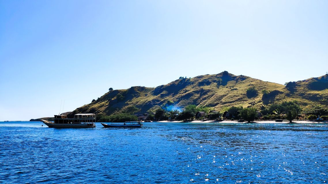 Zwei Boote schwimmen auf glitzernd blauem Wasser vor einer hügeligen grünen Insel mit einer kleinen Siedlung an ihrer Küste.