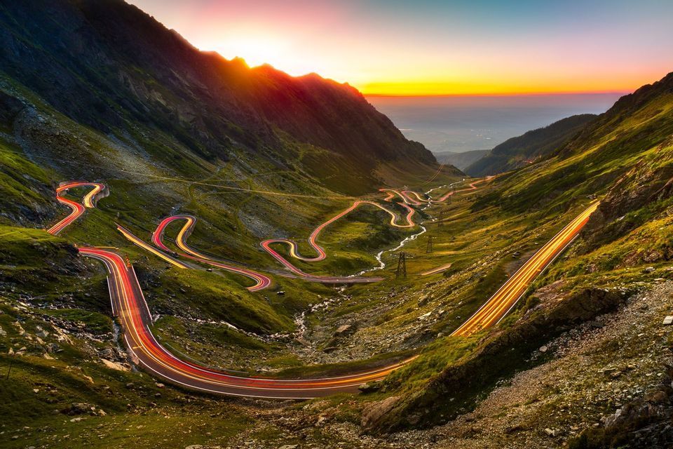 Las estelas de luz de los coches iluminan una carretera sinuosa a través de un puerto de montaña cubierto de hierba mientras el sol se pone en el horizonte.