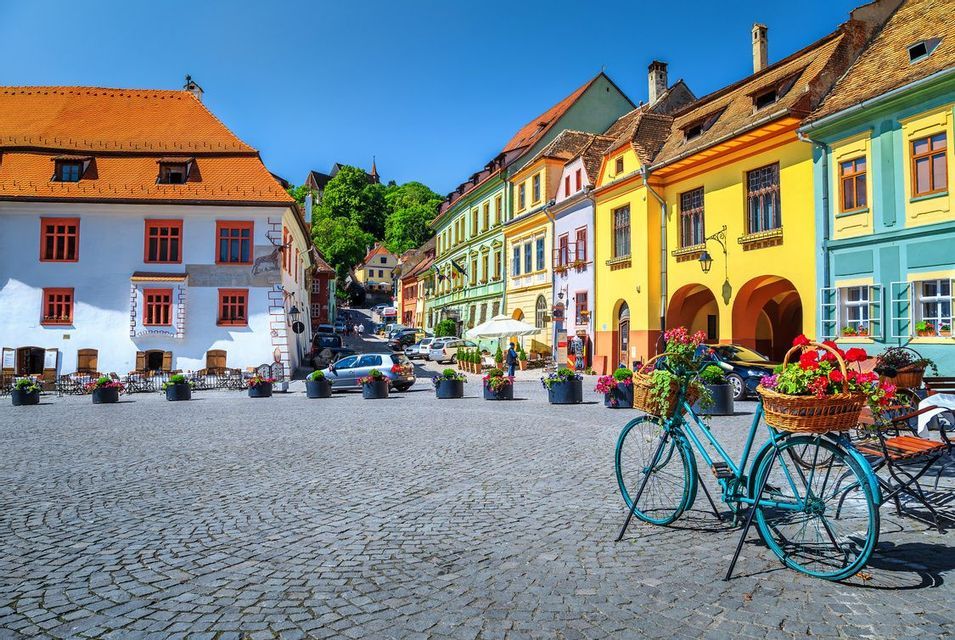 An einem sonnigen Tag steht ein blaues Fahrrad mit Blumenkörben auf einem gepflasterten Platz vor farbenfrohen Gebäuden.