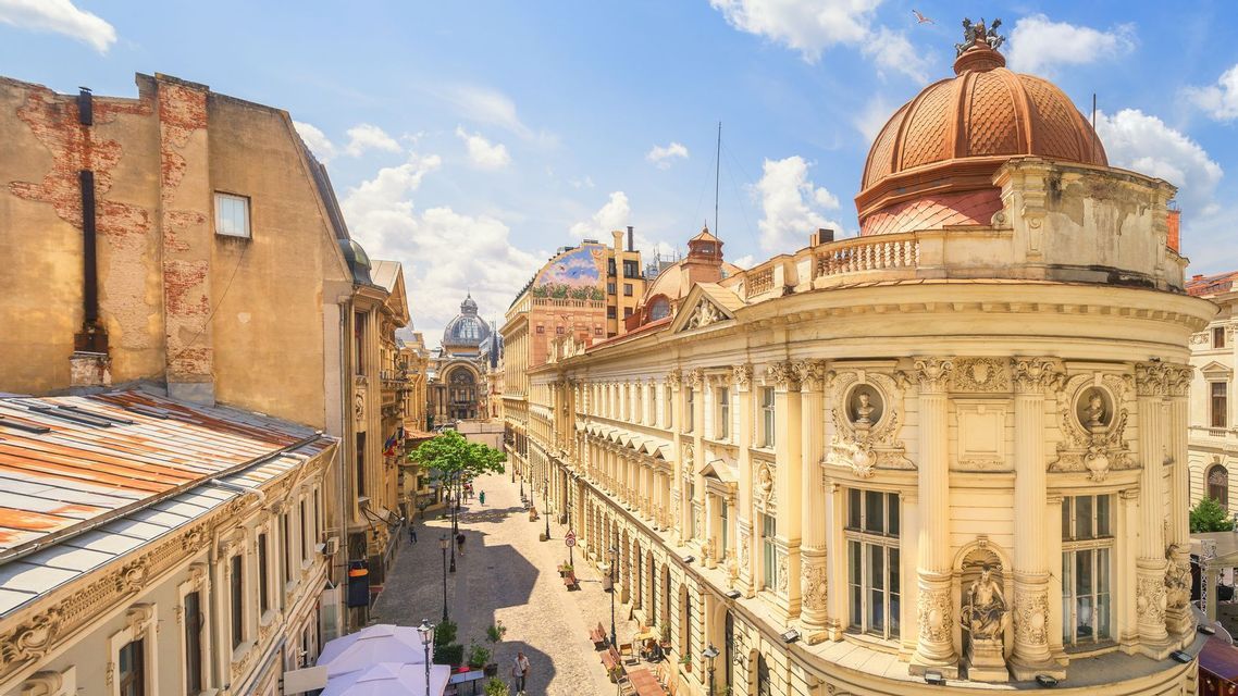 Vue plongeante sur une rue de ville ensoleillée, bordée de bâtiments classiques ornés et dominée par un grand dôme orange sous un ciel bleu.