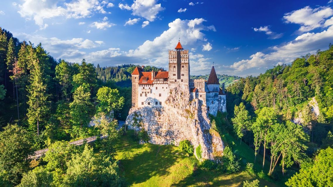 Un castillo blanco con tejados rojos se asienta sobre un acantilado rocoso rodeado por un denso bosque verde bajo un cielo azul con nubes.
