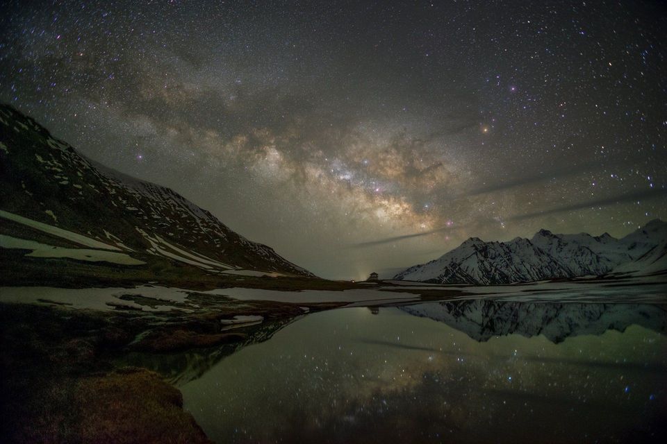 Un cielo notturno stellato con la Via Lattea si riflette in un lago calmo circondato da montagne innevate.