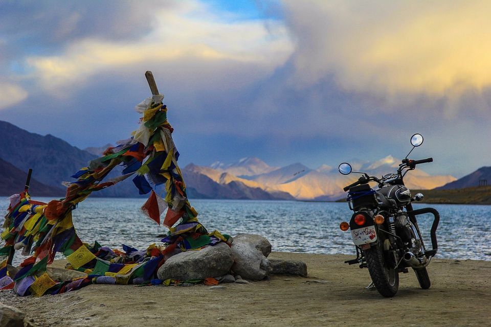 Una moto nera parcheggiata sulla riva di un lago accanto a bandiere di preghiera tibetane colorate, con montagne lontane sotto un cielo nuvoloso.