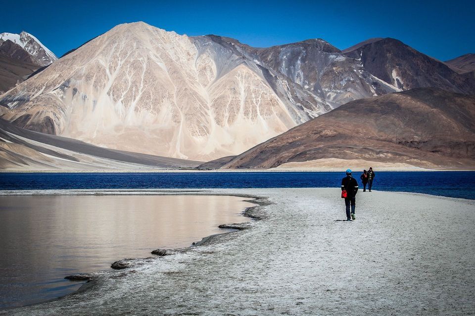 Un viaggio di gruppo WeRoad cammina su una stretta lingua di terra accanto a un lago blu, con grandi montagne aride sullo sfondo.
