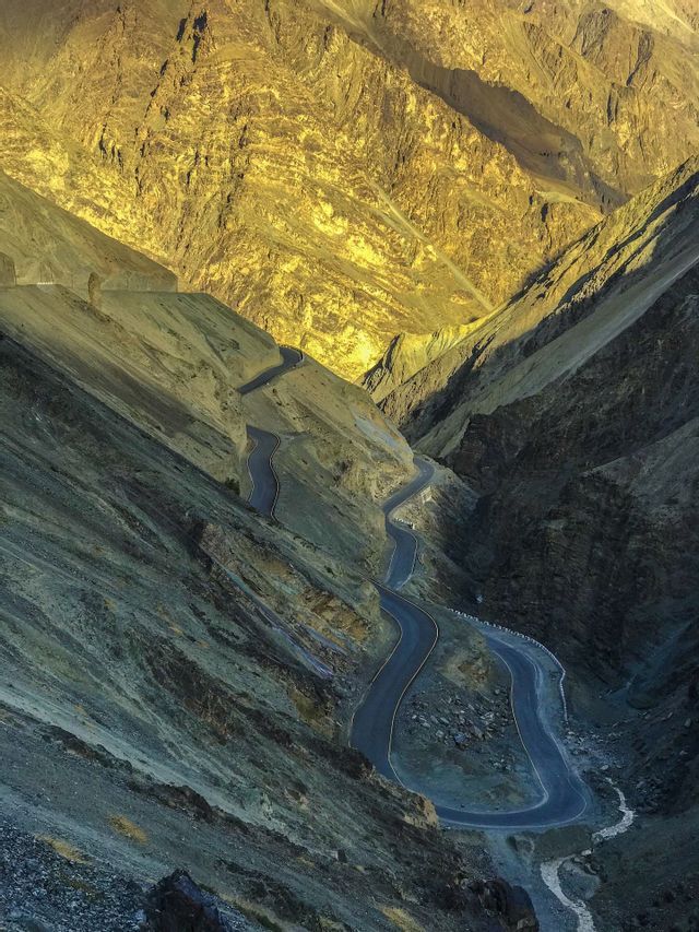 Una strada asfaltata tortuosa si snoda attraverso un profondo passo di montagna con cime illuminate dalla luce dorata del sole.