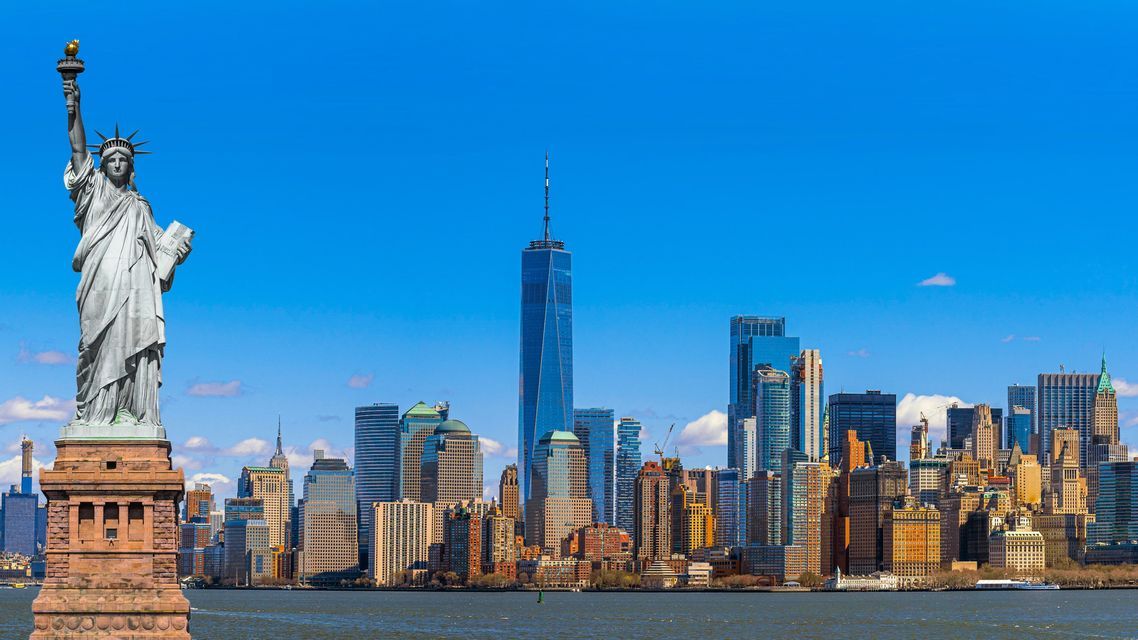 The Statue of Liberty stands before the Manhattan skyline, including the One World Trade Center, under a bright blue sky.