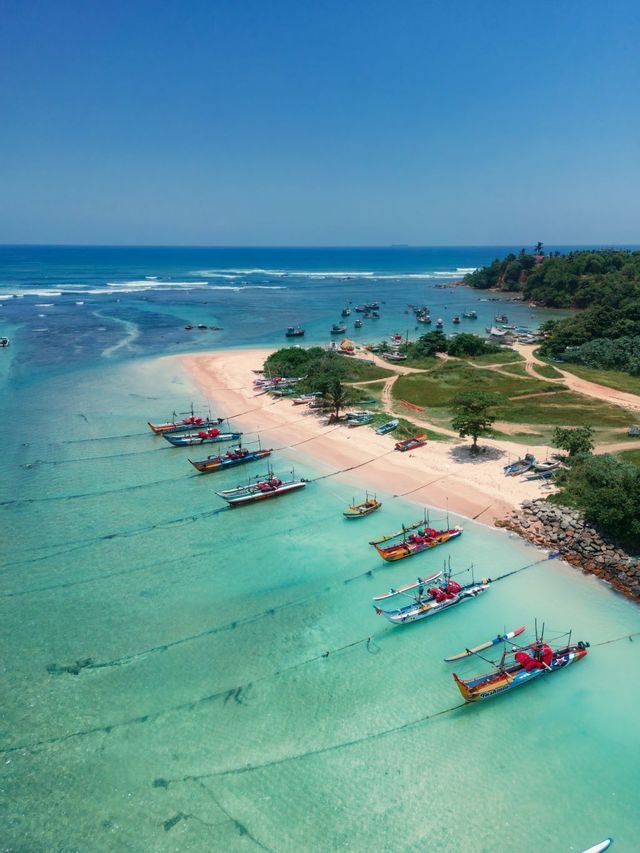 An aerial view of colorful fishing boats moored in a line in shallow, turquoise water next to a sandy beach.