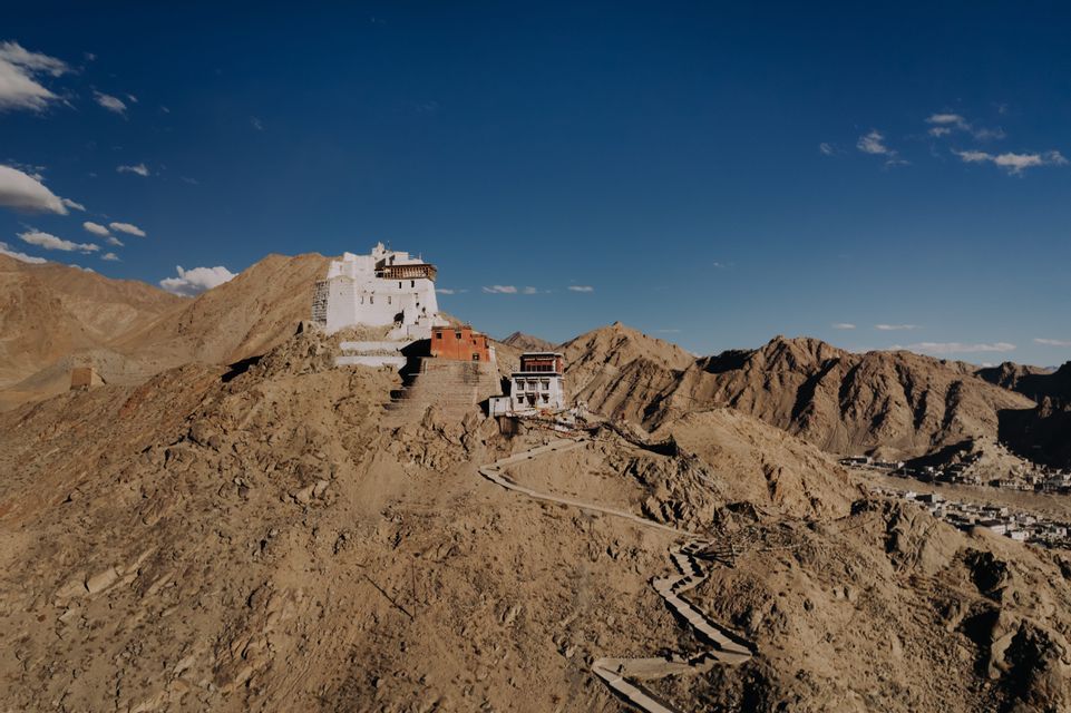 Un complesso monastico bianco si erge in cima a una montagna rocciosa e bruna, con un sentiero tortuoso in pietra che vi conduce sotto un cielo azzurro e limpido.