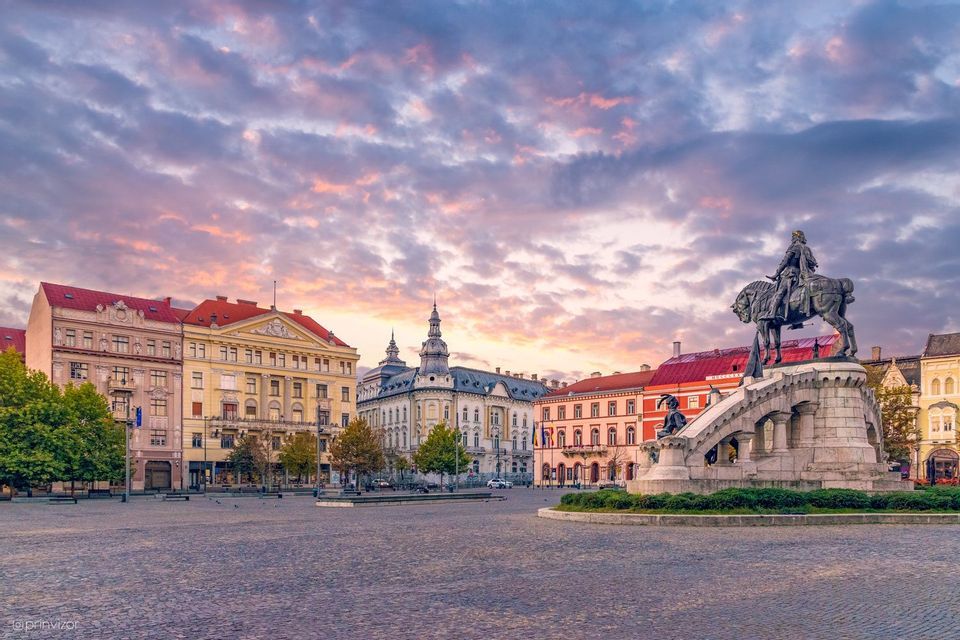 Una statua equestre si erge in una grande piazza acciottolata, circondata da edifici storici sotto un cielo colorato e nuvoloso al tramonto.