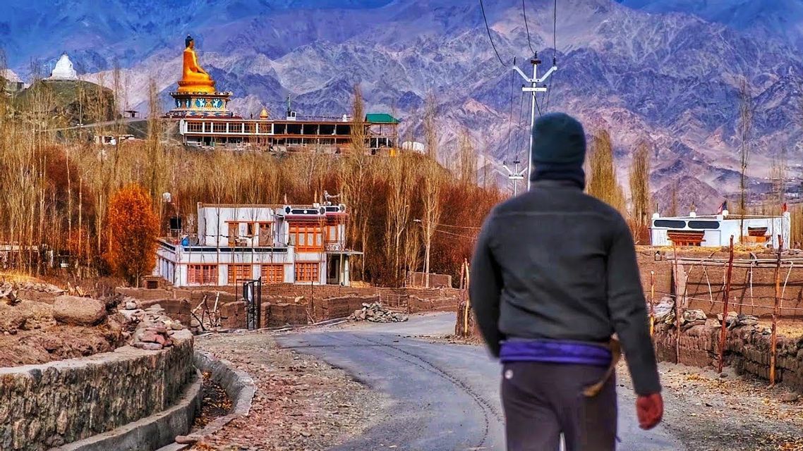 Una persona vista di spalle cammina su una strada verso un villaggio con una grande statua dorata del Buddha, sullo sfondo di una montagna.