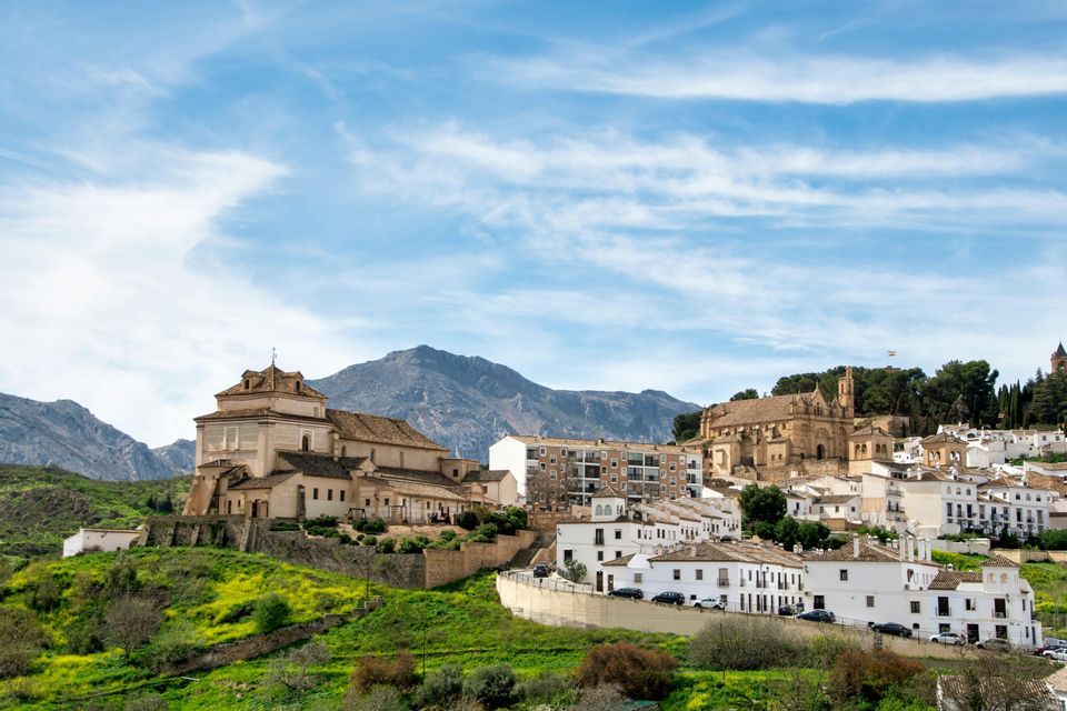 A town of white buildings and stone churches on a lush green hillside, with large mountains in the background under a blue sky.