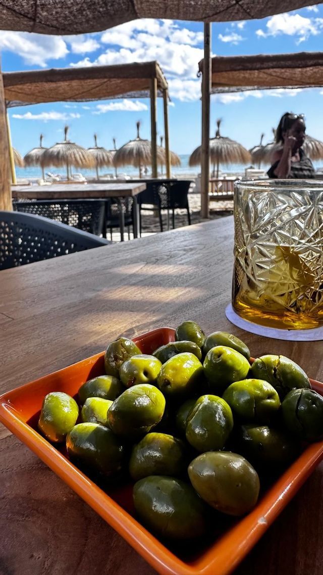 A bowl of green olives and a glass of a beverage on a wooden bar overlooking a sunny beach with thatched umbrellas.