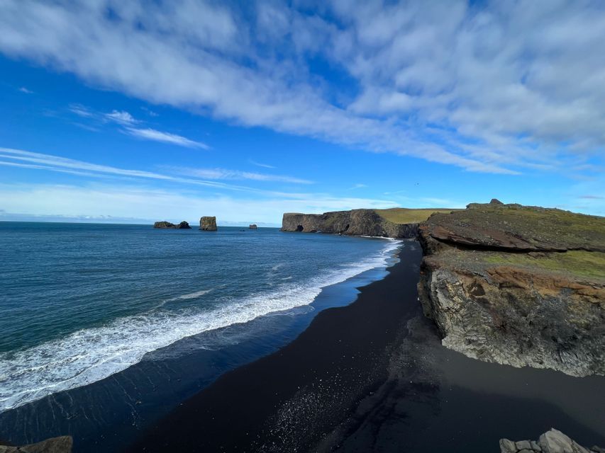 Una vista elevada de una costa con una playa de arena negra, agua azul oscuro y acantilados rocosos bajo un cielo azul con nubes dispersas.
