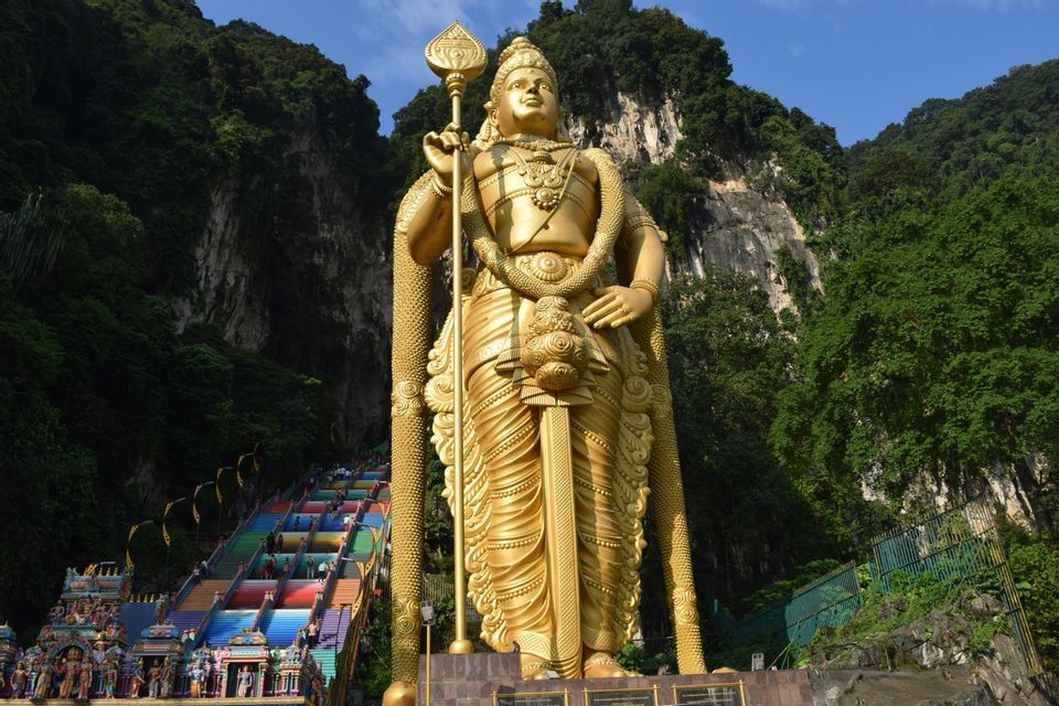 Eine große, goldene Statue einer Hindu-Gottheit steht am Fuße einer Klippe neben einer langen, regenbogenfarbenen Treppe.