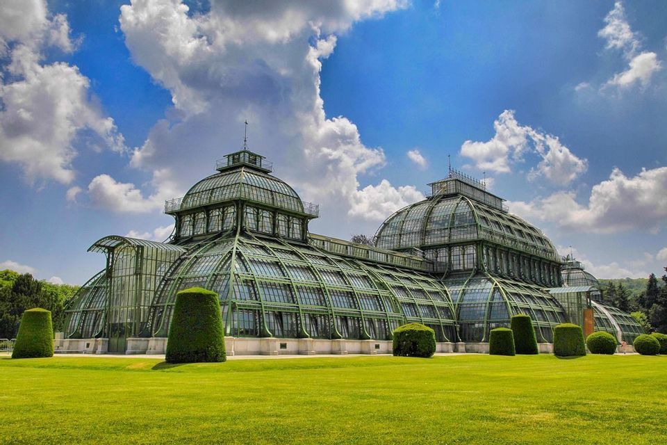 An ornate, glass-paneled greenhouse with a green frame and domes sits on a manicured lawn under a blue sky with white clouds.