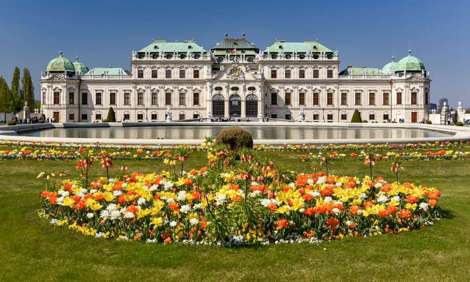 A large baroque palace with green domes seen across a reflecting pool and a lawn with colorful flowerbeds.