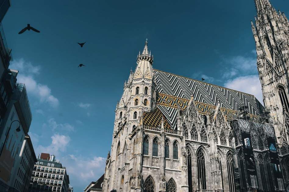 Eine reich verzierte gotische Kathedrale mit einem farbenfrohen, geometrisch gemusterten Ziegeldach, von unten vor blauem Himmel mit Vögeln gesehen.