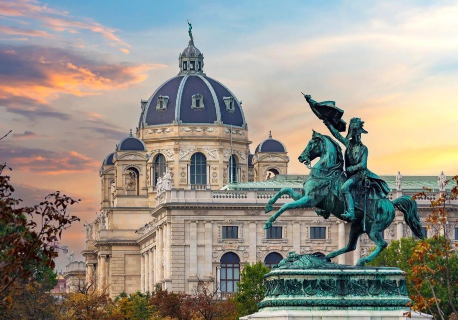 Una statua equestre in bronzo su un piedistallo di fronte a un grande edificio ornato con una cupola prominente sotto un cielo al tramonto.