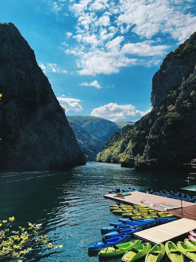 Kayak colorati si trovano su un molo di legno lungo un fiume che scorre attraverso un canyon ripido e alberato sotto un cielo azzurro e nuvoloso.