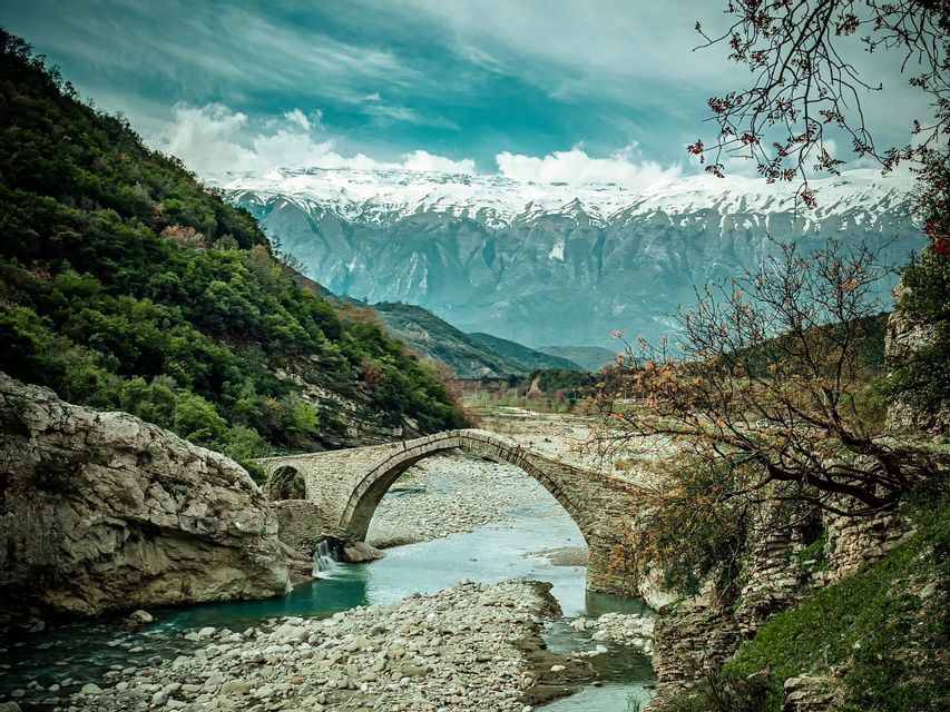 Eine Steinbogenbrücke überquert einen Fluss in einem felsigen Tal, mit imposanten, schneebedeckten Bergen im Hintergrund.