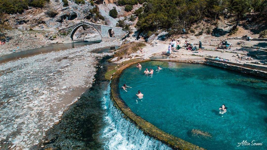 Vue aérienne d'un groupe WeRoad en voyage se baignant dans une piscine thermale turquoise, avec un pont en arc de pierre et une rivière en arrière-plan.