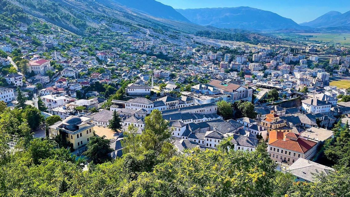 Veduta aerea di una città fitta con tetti in pietra ed edifici bianchi, sullo sfondo di dolci montagne verdi.