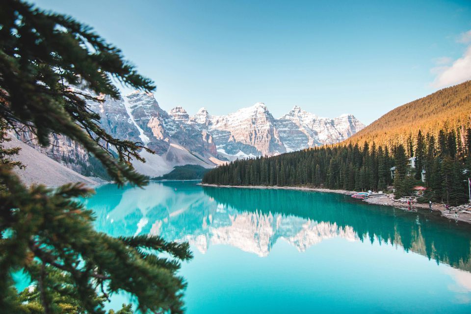 Montagnes enneigées et forêt de pins dense se reflétant dans l'eau calme et turquoise d'un lac sous un ciel clair.
