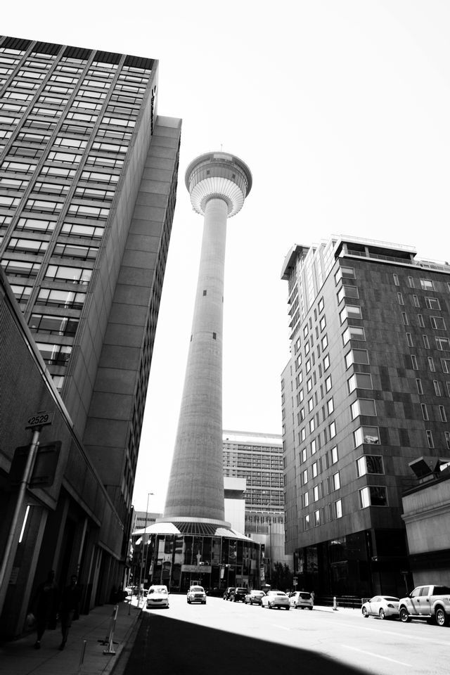 Une vue en noir et blanc, en contre-plongée, d'une haute tour d'observation encadrée par des gratte-ciel modernes dans une rue de ville.