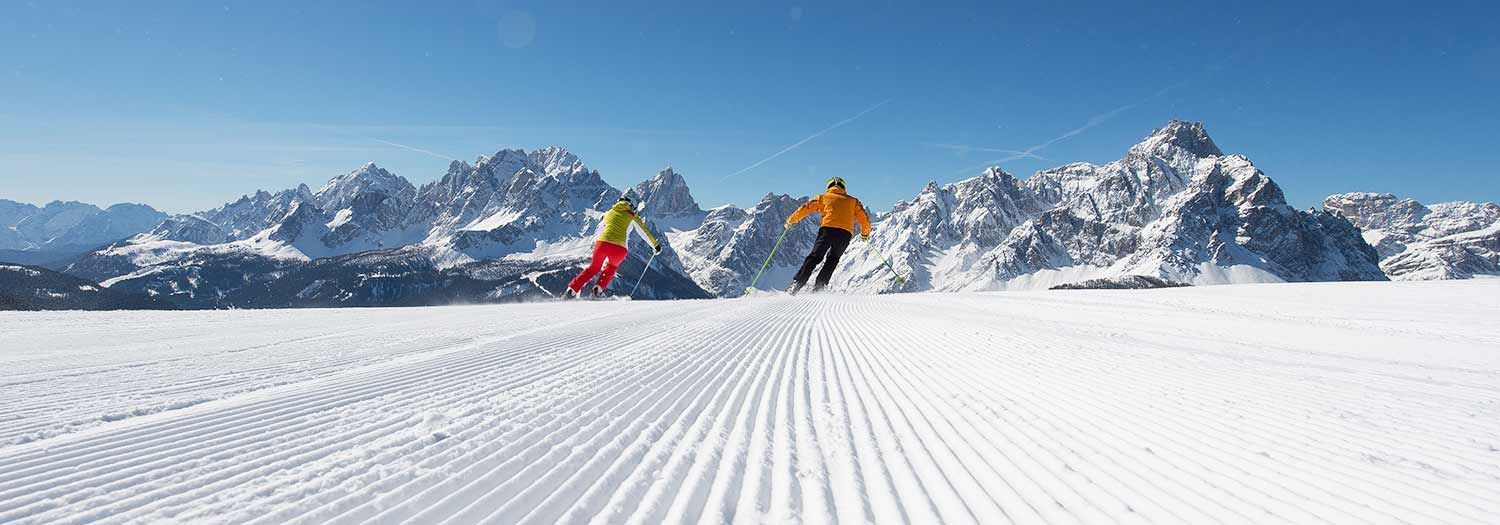 Zwei Skifahrer in bunten Jacken fahren eine präparierte Skipiste hinab mit Panoramablick auf eine Bergkette unter klarem Himmel.