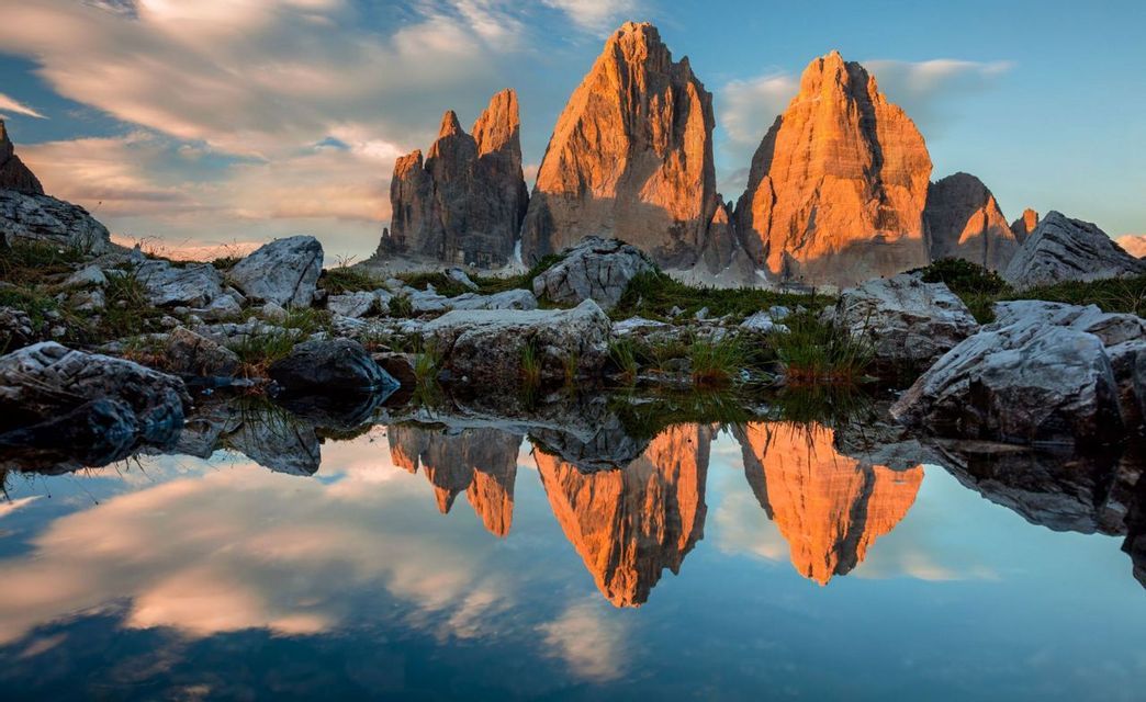Gezackte Berggipfel, beleuchtet von goldenem Licht, spiegeln sich in einem stillen See mit felsigem Ufer im Vordergrund.