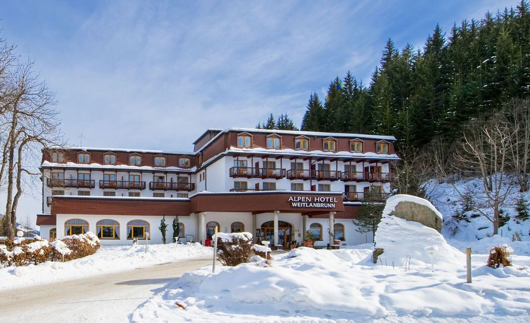 Ein großes, mehrstöckiges Alpenhotel liegt in einer verschneiten Landschaft am Fuße eines Kiefernwaldes unter einem teils bewölkten Himmel.
